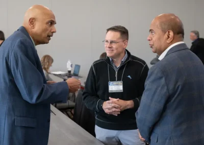 Three professionals in business attire have a conversation during a networking event, with other attendees visible in the background.