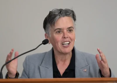A speaker in a gray blazer addresses an audience from a podium, with an SIUE pin visible on her lapel.