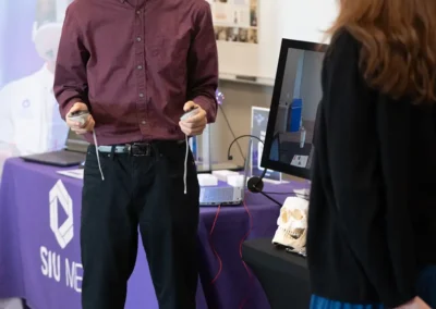 A student tries a VR headset at an SIU Medicine immersive learning demonstration booth, with a human skull model and monitor visible on the table behind him.