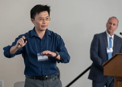 A presenter wearing a conference badge addresses an audience in a meeting room, with another person standing near a podium in the background.