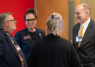 Four professionals wearing name badges have a conversation in a hallway during a conference or networking event.