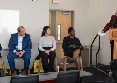 A moderator in a red blazer introduces a panel of four speakers seated on a stage at a Southern Illinois University Edwardsville event.